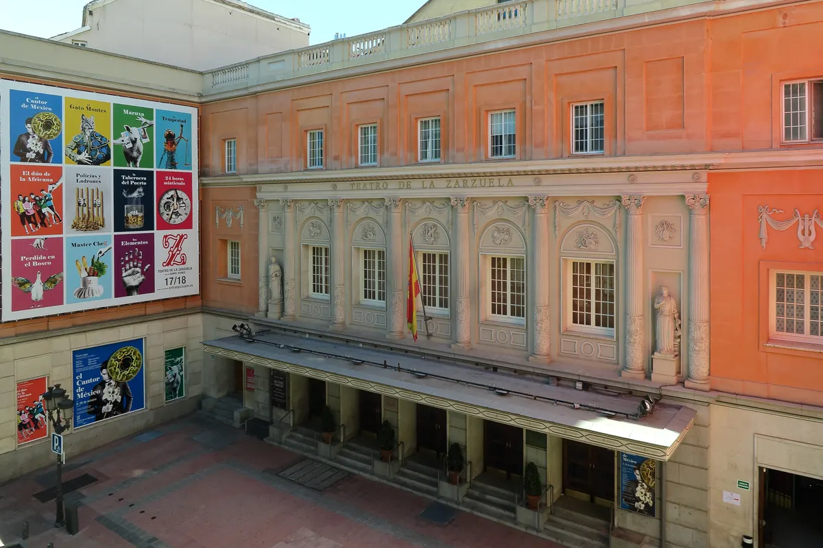 The pink and cream stone front of the Teatro de la Zarzuela, seen from a courtyard with posters on a wall.