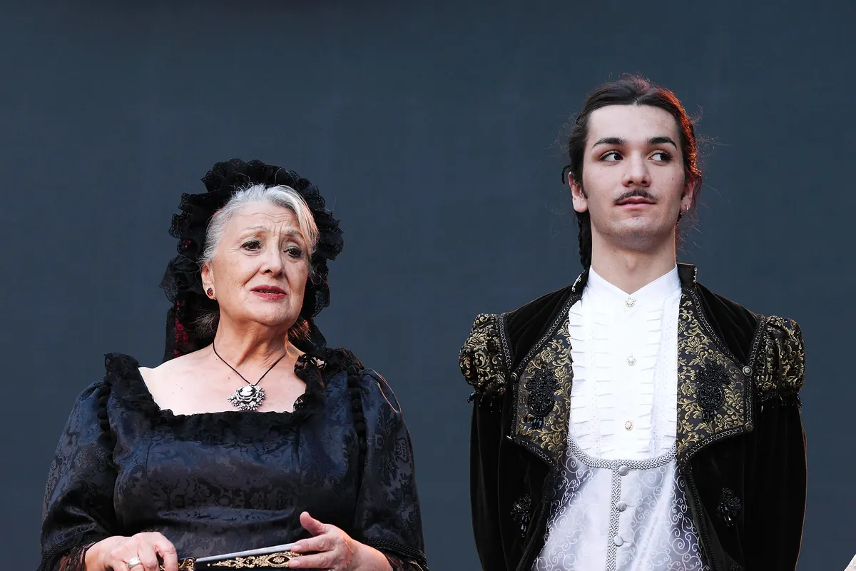 A woman in a white lace headdress holding a fan, a young man in a dark embroidered jacket and a woman in a cream blouse stand in stage light