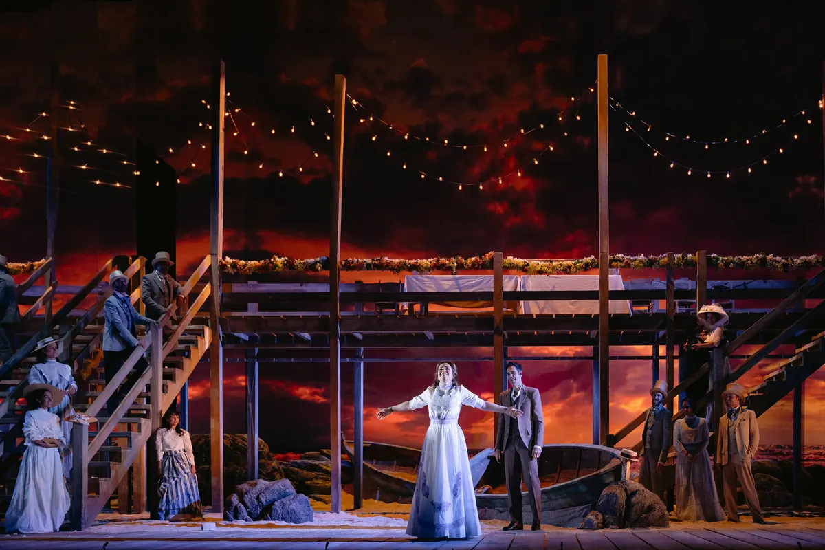 Two singers on a stage set with a red and gold sunset sky, a wooden jetty and strings of lights.